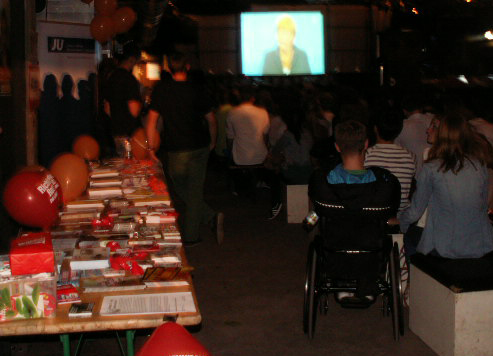 Das Bild zeigt die Halle 02 in Heidelberg am Sonntag, den 01.09.2013. Im Intergrund ist Angela Merkel auf einer großen Leinwand zu sehen. Das ist die Übertragung des TV-Duells mit Peer Steinbrück. Hinter den Stuhlreihen ist ein Rollstuhlfahrer von hinten zu sehen. Links daneben ist ein Parteistand mit vielen roten Luftballons und Infomaterial. Da alle Stühle voll besetzt sind, stehen auch einige Leute.